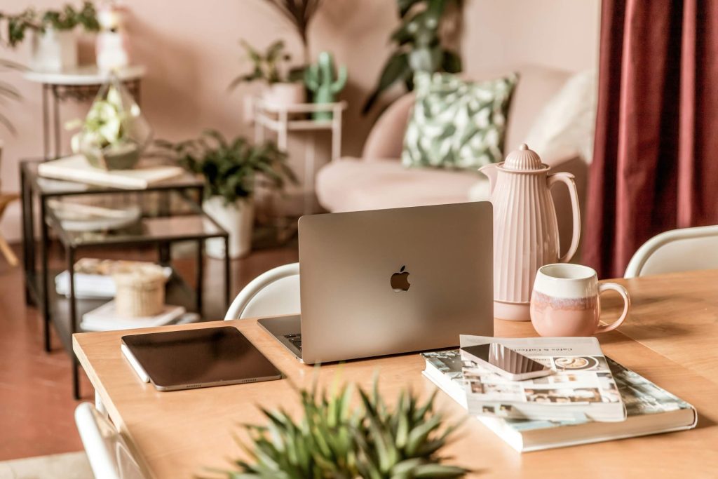 Lap top on a table, next to an ipad. With a soft focus background of a chair and side tables. All in pink hues
